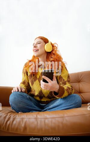 Une femme joyeuse aux cheveux roux écoute de la musique à travers des écouteurs jaunes tout en étant assise les jambes croisées sur un canapé en cuir marron, tenant un smartphone et Banque D'Images