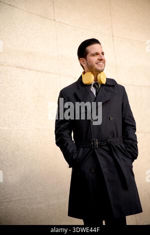 Homme d'affaires dans un trench-coat sombre, debout à l'extérieur contre un mur texturé, sourit en toute confiance tout en portant de grands écouteurs jaunes autour de son cou Banque D'Images