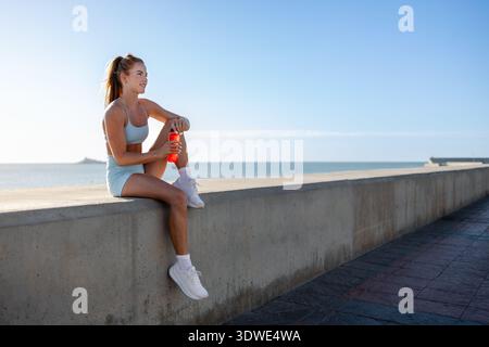 Une femme en tenue athlétique est assise sur un mur de bord de mer à Malaga, en Espagne. Après sa course, elle se repose avec une bouteille d'eau rouge vif, profitant de la vue sur l'océan et Banque D'Images