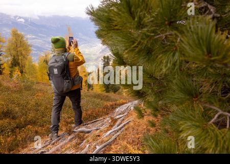 Un randonneur dans un chapeau vert vif et sac à dos explore le paysage d'automne à couper le souffle des Alpes, capturant la vue panoramique des forêts colorées A. Banque D'Images