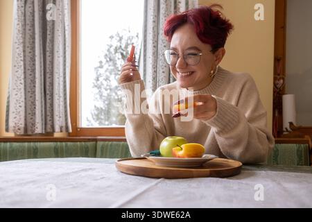 Une jeune femme joyeuse est assise à une table dans un environnement familial confortable, appréciant joyeusement les fruits et légumes frais, mettant en valeur une vie saine et joyeuse Banque D'Images
