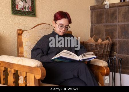 Une jeune femme aux cheveux rouges courts et aux lunettes est confortablement assise dans un fauteuil, profondément immergée dans un livre. L'ambiance chaleureuse de sa maison ajoute à la s. Banque D'Images