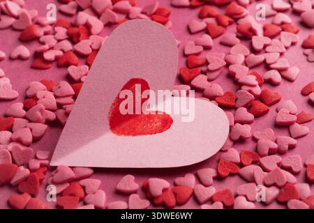 Un coeur en papier rose avec un motif de coeur d'empreinte de doigt rouge repose au milieu de cœurs de confettis de sucre rouge et rose dispersés sur un fond assorti, évoquant Valen Banque D'Images