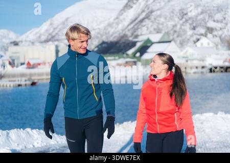 Un couple joyeux se promène dans le pittoresque paysage hivernal des Lofoten. Les montagnes enneigées et un fjord serein créent une toile de fond idyllique à leur Banque D'Images