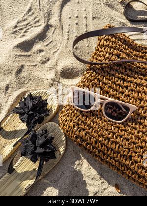 Des lunettes de soleil élégantes reposent sur un sac de plage tissé aux côtés de tongs fleuries sur le sable. Cette scène estivale évoque une journée de détente à la plage, se mélangeant Banque D'Images