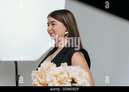 Une femme pose en toute confiance dans un cadre de studio, tenant un bouquet de fleurs beiges. L'éclairage doux et le fond minimaliste soulignent son éléganc Banque D'Images