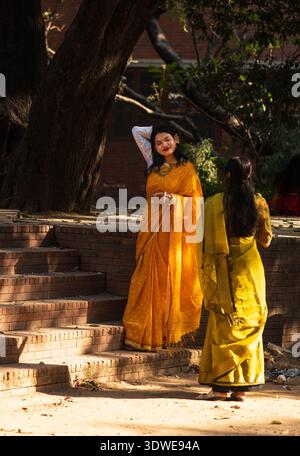 Dhaka, Bangladesh - 14 février 2026 : vue d'une femme dans un sari jaune vif se tient sur des marches ensoleillées près d'un grand arbre, projetant des ombres et des illuminations Banque D'Images