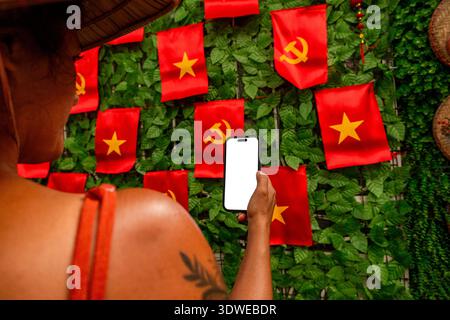 Tourist tient un smartphone avec un écran vierge, capturant de petits drapeaux rouges avec une étoile jaune et des symboles marteau et faucille sur un mur verdoyant Banque D'Images