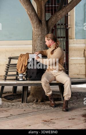 Un homme vêtu de façon décontractée avec une queue de cheval est assis sur un banc à l'extérieur. Il tend la main dans un sac à dos noir pour récupérer un ordinateur portable, avec une tasse de café ne Banque D'Images