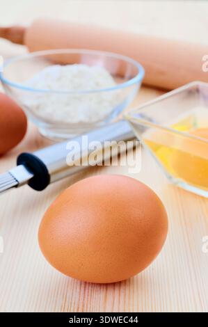 Une image en gros plan présentant des ingrédients essentiels à la cuisson, y compris un œuf brun, un bol de farine et un petit bol d'œufs cassés sur une table en bois, Banque D'Images