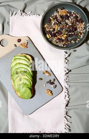 Un délicieux petit déjeuner sain de pain grillé à l'avocat garni de graines de chia sur une planche à découper en pierre. Un bol de noix et de graines mélangées se trouve à proximité sur un Li Banque D'Images
