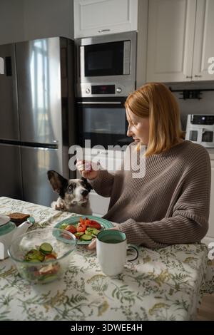 Une femme enceinte s'assoit confortablement à une table de cuisine en dégustant un repas sain. Son chien est assis à côté d'elle, regardant avec impatience qu'elle s'engage dans son ro quotidien Banque D'Images