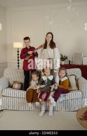 Une famille joyeuse se réunit dans un salon confortable pendant Noël. Les enfants sont habillés festivement, l'un jouant un ukulélé, tous partageant des moments joyeux sur Banque D'Images
