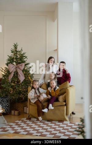 Un joyeux groupe de frères et sœurs assis ensemble dans une pièce confortable, appréciant la saison des fêtes près d'un arbre de noël joliment décoré avec des lumières et des pr Banque D'Images