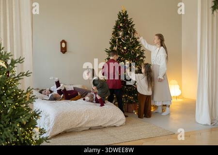 Une famille joyeuse se réunit autour d'un sapin de Noël magnifiquement éclairé, le décorant avec des ornements et des rubans, capturant l'essence de l'esprit de vacances et Banque D'Images