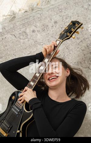 Une femme joyeuse allongée sur un tapis à motifs, enserrant de manière ludique une guitare électrique noire. Son rire incarne un sentiment de détente et de passion pour la musique. Banque D'Images