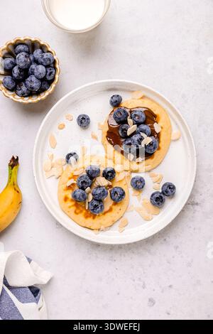 Assiette de petit déjeuner alléchante avec crêpes moelleuses garnies de bleuets frais et d'amandes tranchées, accompagnée d'une banane mûre et d'un verre de mi Banque D'Images