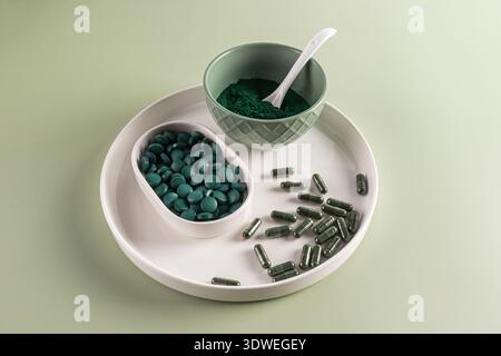Du haut de spiruline dans un bol vert avec une cuillère blanche, des comprimés dans un plat blanc, et des capsules sur un plateau blanc. L'arrière-plan est d'une couleur verte douce, Banque D'Images