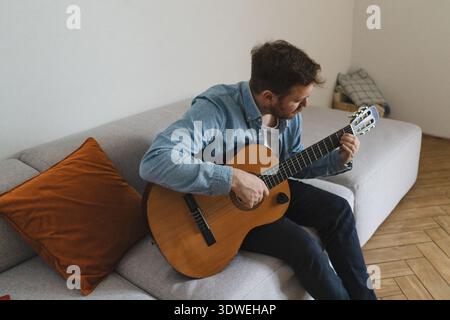 Un jeune homme aime jouer de la guitare acoustique tout en étant assis confortablement sur un canapé confortable à la maison, créant une atmosphère paisible et musicale dans le salon Banque D'Images