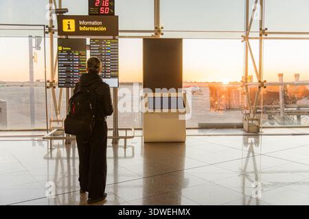 Un voyageur avec un sac à dos se tient à un terminal d'aéroport, vérifiant le tableau de départ. La lumière du soleil tôt le matin filtre à travers les grandes fenêtres, Banque D'Images