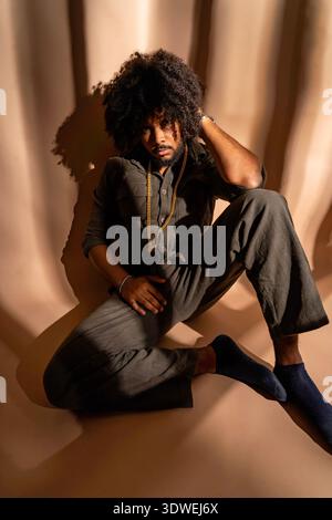 Un homme avec une coiffure afro portant une tenue élégante pose soigneusement dans un cadre de studio bien éclairé. Le jeu des ombres et de la lumière ajoute de la profondeur au Banque D'Images