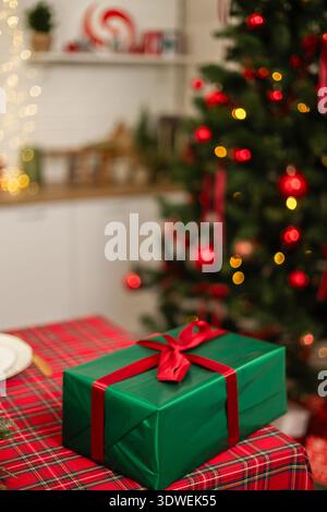 Un cadeau vert magnifiquement enveloppé avec un ruban rouge se trouve sur une nappe à carreaux devant un sapin de Noël brillant orné d'ornements rouges et chaleureux Banque D'Images