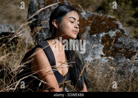 Jeune femme routard prend une pause de la randonnée, assis parmi les hautes herbes et profiter de la lumière chaude du soleil sur son visage, avec un terrain rocheux dans le b. Banque D'Images