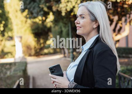 Femme d'affaires senior portant une veste noire et une chemise bleue tient un smartphone et regarde loin tout en pensant à quelque chose dans un parc pendant un Banque D'Images