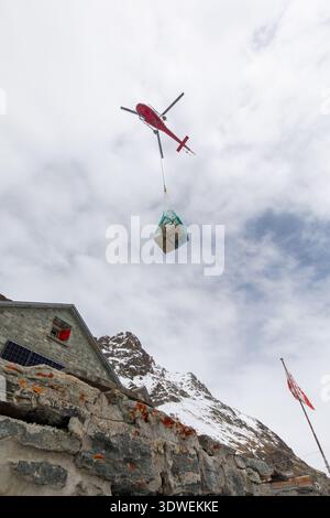 D'en bas, un hélicoptère rouge transporte des fournitures au-dessus d'un sommet enneigé jusqu'à une cabane de montagne isolée à Schonbiel, Zermatt, Valais. Un drapeau suisse agite ami Banque D'Images