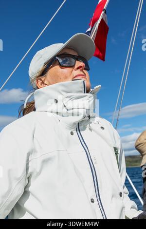 Une femme navigue sur un voilier par temps clair en Norvège, enfilant des lunettes de soleil et une casquette. Le drapeau norvégien bat en arrière-plan contre un bl lumineux Banque D'Images