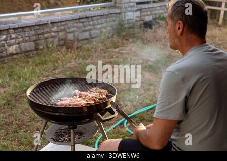 Un homme fait cuire des brochettes de poulet sur un grill au charbon de bois dans son arrière-cour, profitant d'un barbecue relaxant au coucher du soleil. Le cadre extérieur paisible est parfait Banque D'Images