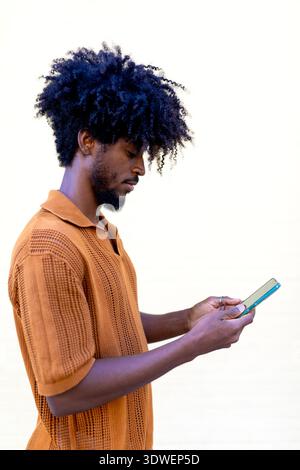 Homme noir aux cheveux bouclés debout sur un fond clair, concentré sur son smartphone. Il semble engagé et réfléchi, portant un s orange décontracté Banque D'Images