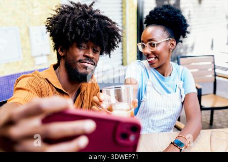 Un couple joyeux assis dehors, partageant un verre et capturant le moment avec un selfie. Ils sourient, apprécient la compagnie de l'autre sur un d ensoleillé Banque D'Images