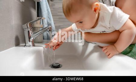 Un bébé se lave les mains avec les conseils de ses parents. L'eau coule doucement du robinet que la famille favorise l'hygiène et le lien dans un e nourrissant Banque D'Images