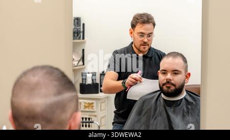 Un barbier se prépare à donner une coupe de cheveux dans un salon de coiffure moderne. Le client est assis dans la chaise avec un regard concentré que le coiffeur se prépare avec le sien aussi Banque D'Images