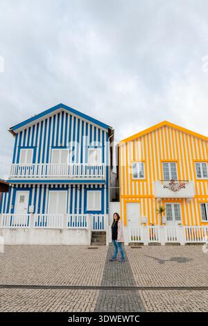 Une personne se tient devant les maisons rayées vibrantes et colorées de Costa Nova. L'architecture charmante présente des rayures alternées en bleu et Banque D'Images