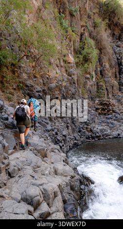 Une touriste suit un guide Massaï le long d'un sentier rocheux menant à la cascade Engare Sero. Ils explorent le magnifique paysage naturel entouré Banque D'Images
