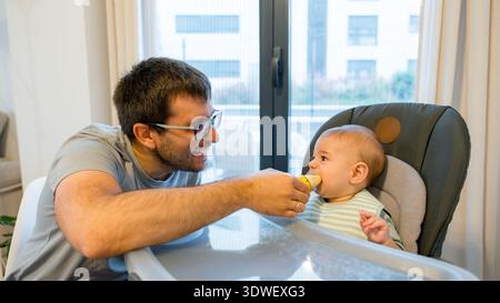 Un parent aimant nourrit son bébé d'une banane dans le cadre du sevrage mené par bébé. Le bébé, assis dans une chaise haute, explore le goût et la texture du fruit, Banque D'Images
