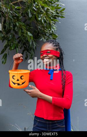 Adolescente afro-américaine posant au mur gris, portant un masque rouge, une cape bleue, tenant un seau orange Banque D'Images