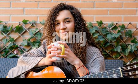 Une jeune femme aux cheveux bouclés profite d'un moment de détente, tenant une tasse tout en étant assise avec une guitare. Elle respire le calme et le contentement dans un outd confortable Banque D'Images