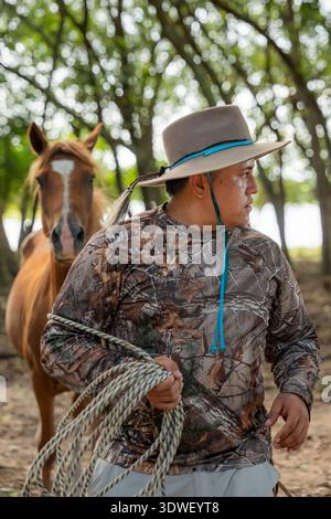 Un cow-boy colombien à Casanare, en Colombie, se dresse dans une forêt luxuriante avec un cheval à proximité. Il porte un chapeau à larges bords et des vêtements de camouflage, tenant un Banque D'Images