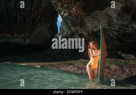 Une femme en maillot de bain tenant une planche de surf se tient dans les eaux claires d'une grotte à Bali. Le soleil brille sur elle, soulignant le beau serein et exotique Banque D'Images