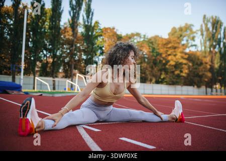 L'athlète s'étire sur une piste de stade, se préparant à l'entraînement. Portant un équipement sportif, elle fait preuve de concentration et de flexibilité dans un set de fitness extérieur Banque D'Images