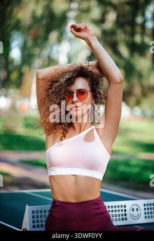 Une athlète confiante portant des vêtements de sport élégants et des lunettes de soleil tendance étire ses bras à l'extérieur. Entourée de verdure, elle incarne le fitness et le bien-être Banque D'Images