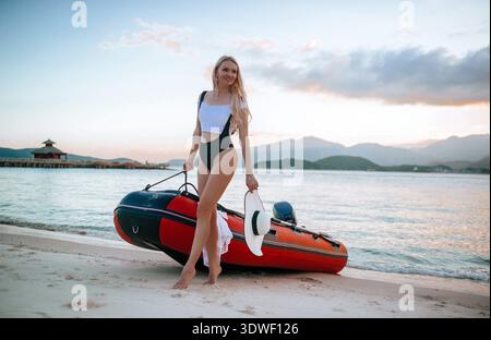 Une femme dans un maillot de bain élégant pose avec un bateau gonflable sur les rives sablonneuses de l'île de Vinpearl, Nha Trang, Vietnam, capturant une scène de coucher de soleil sereine Banque D'Images