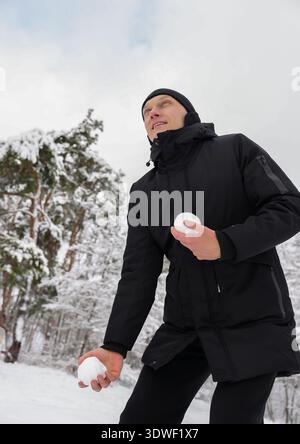 Un homme portant un manteau d'hiver noir tient joyeusement des boules de neige dans un paysage enneigé, indiquant une atmosphère hivernale ludique et joyeuse. Banque D'Images
