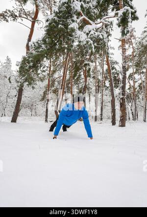 Un homme fait de l'exercice en plein air dans une forêt enneigée, faisant preuve de détermination et de résilience. Portant une veste bleue et un bonnet noir, il souligne l'imp Banque D'Images