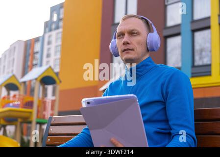 Un homme en pull bleu est assis sur un banc extérieur, tenant sa tablette et portant des écouteurs. Le bâtiment coloré en arrière-plan suggère un vibran Banque D'Images