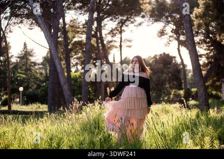 Une jeune femme enceinte dans une jupe fluide marche gracieusement à travers une forêt ensoleillée, entourée de grands arbres et de verdure luxuriante, incarnant la tranquillité Banque D'Images