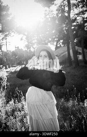 Une jeune femme enceinte se tient gracieusement dans un cadre naturel ensoleillé, portant une jupe fluide et un chapeau. Son expression sereine incarne la beauté de Mo Banque D'Images